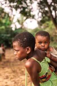 A young African child carries a baby sibling in a serene countryside setting, capturing a moment of familial support.
