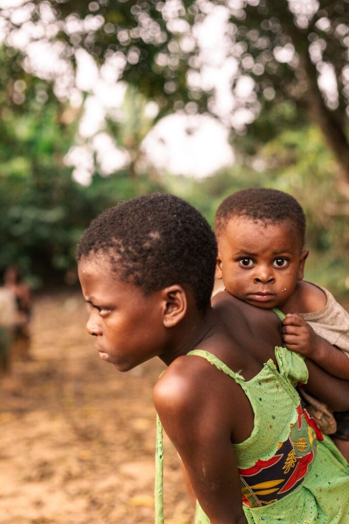 A young African child carries a baby sibling in a serene countryside setting, capturing a moment of familial support.